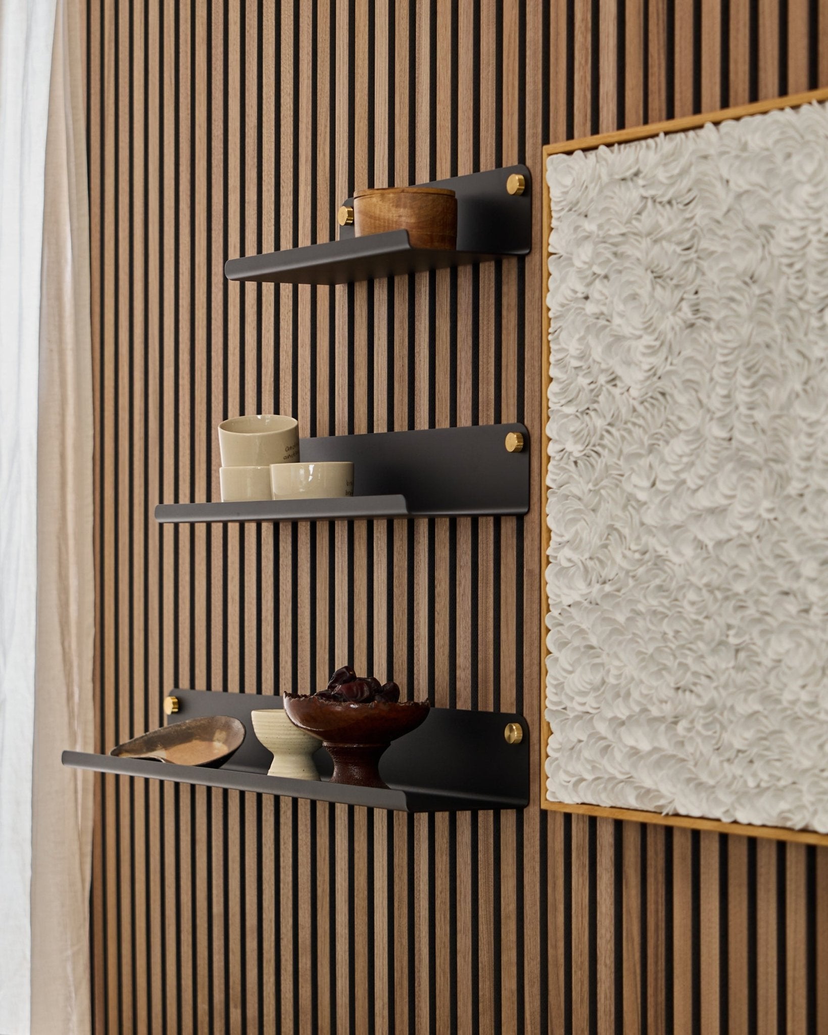 Tres baldas anchas WoodUpp Norr black (51,9 cm) montadas sobre un panel acústico de madera WoodUpp Akupanel, junto a una obra de arte blanca con textura, con cuencos y recipientes de madera expuestos.